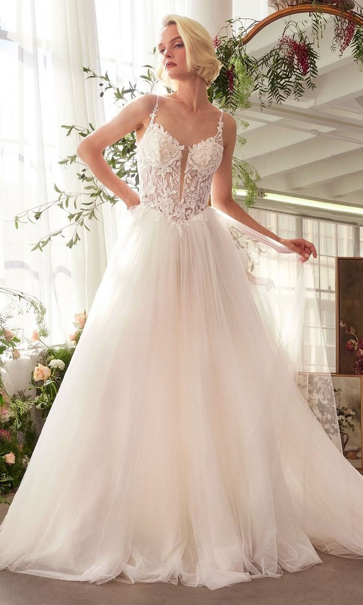 A woman stands indoors near flowers and a mirror, holding part of her skirt while wearing the Andrea and Leo WL029 long wedding dress—a sleeveless gown with a lace corset bodice and an A-line tulle skirt.